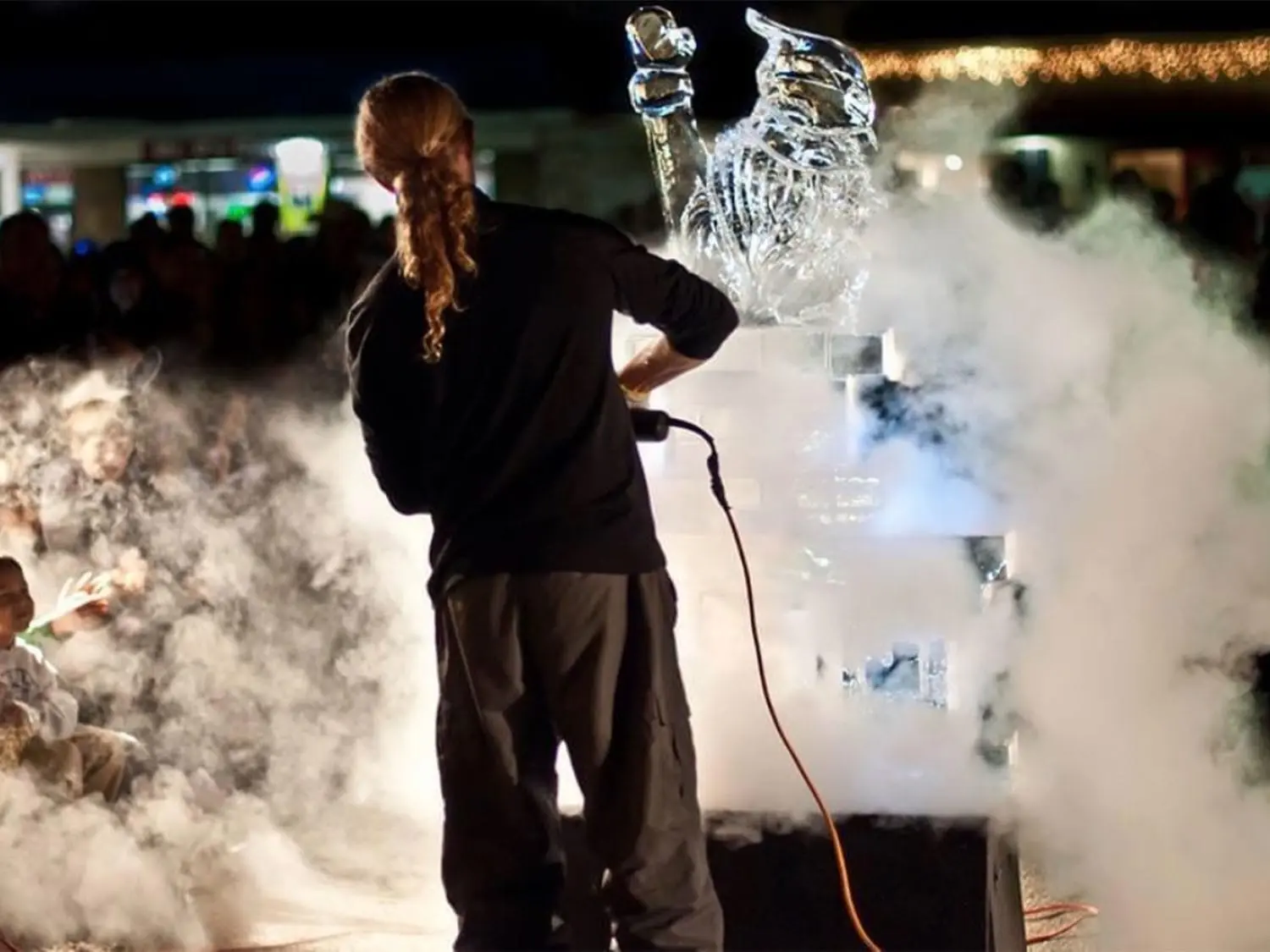 Master ice sculptor Buddy Rasmussen skillfully carves an intricate design from a large ice block during an outdoor event.